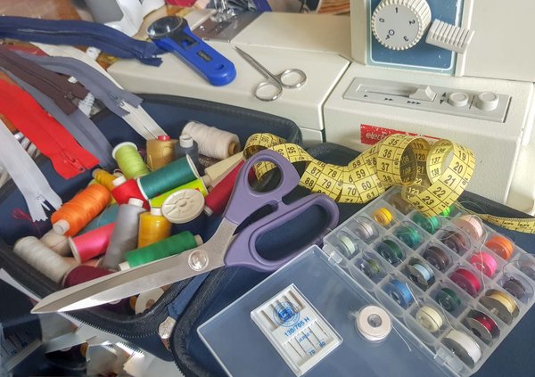 Reißverschlüsse und Garn in verschiedenen Liegen durcheinander vor einer Nähmaschine. Dazu liegt noch ein Maßband und eine Schere mit Lila Griff .
